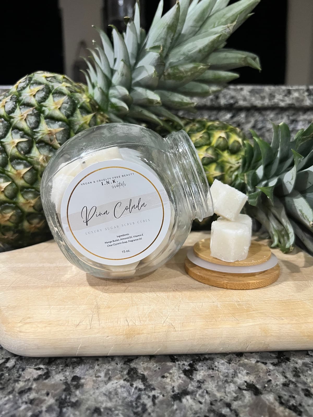 A jar of I.N.K. essentials Piña Colada luxury sugar scrub cubes tipped on its side, with two cubes resting on the lid beside fresh pineapple